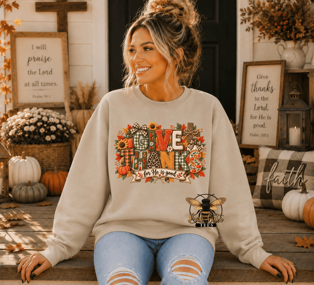 A woman wearing a beige crewneck sweatshirt with a colorful 'GIVE THANKS' autumn graphic sits on a wooden porch. The graphic features pumpkins, fall leaves, and a bee logo.