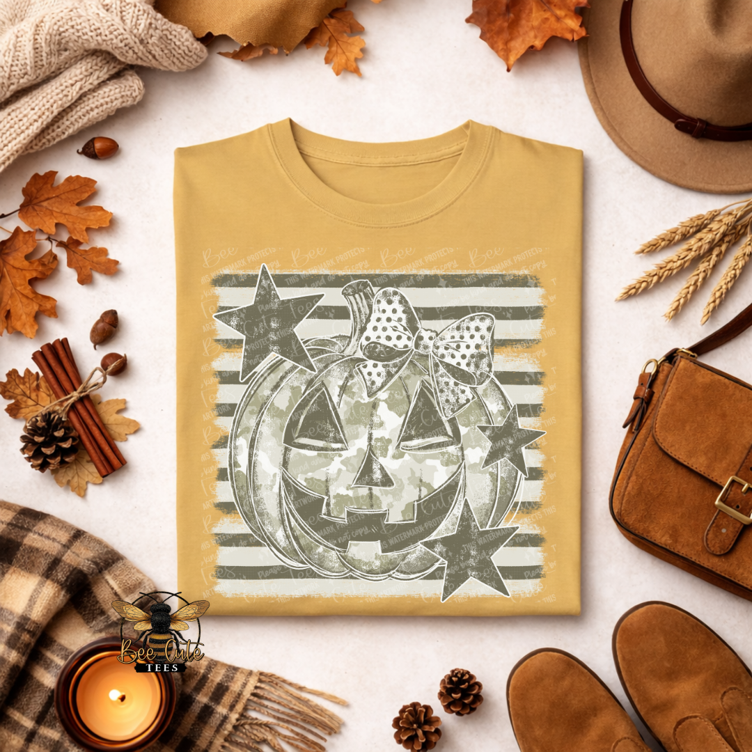 A folded tan crewneck t-shirt with a Halloween graphic of a jack-o'-lantern in camouflage, surrounded by autumn flat-lay props including a hat, shoes, a candle, and leaves on a light background.