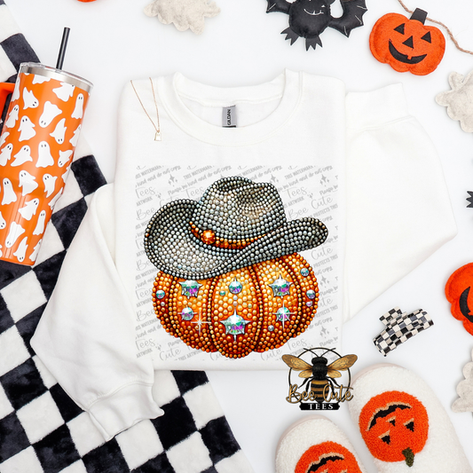 A white long-sleeve sweatshirt laid flat on a white background, decorated with a large Halloween graphic of an orange pumpkin covered in rhinestones and a black cowboy hat. The sweatshirt is surrounded by Halloween-themed props including a black-and-white checkered mat, an orange tumbler with ghost prints, jack-o'-lantern decorations, and fuzzy slippers.
