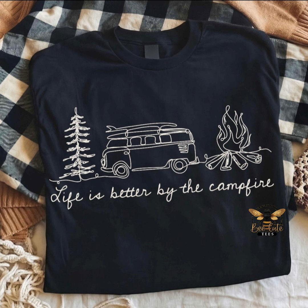 A navy blue t-shirt with a white line art graphic of a camp van, trees, and a campfire, with the text 'Life is better by the campfire'. The shirt is laid flat on a black and white buffalo plaid blanket with cozy knitted blankets in the background.