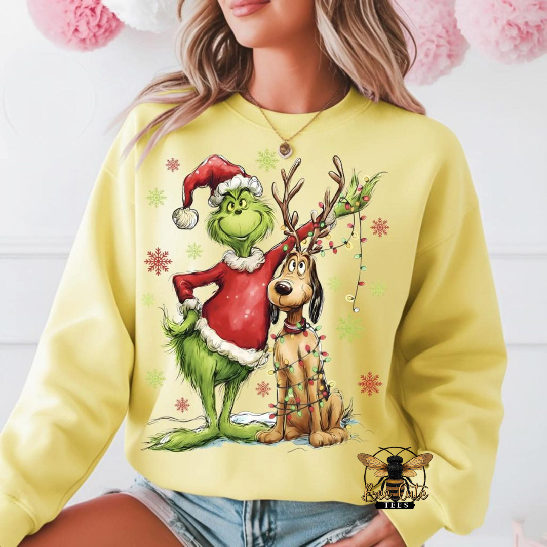 A woman wearing a pale yellow sweatshirt with a large graphic of a character and a dog dressed as Santa, with holiday decorations and snowflakes. The photo is cropped from the shoulders to the hips, with a white paneled wall and pink pom-pom decorations in the background.