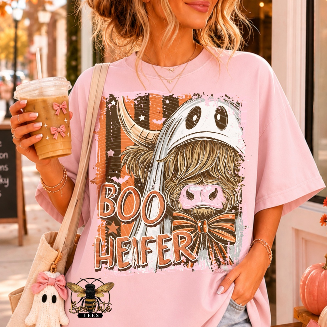 A woman wearing a light pink graphic t-shirt with a cartoon ghost pig design and the text 'BOO HEFER'. She is holding an iced coffee and wearing a beige tote bag in a storefront setting.