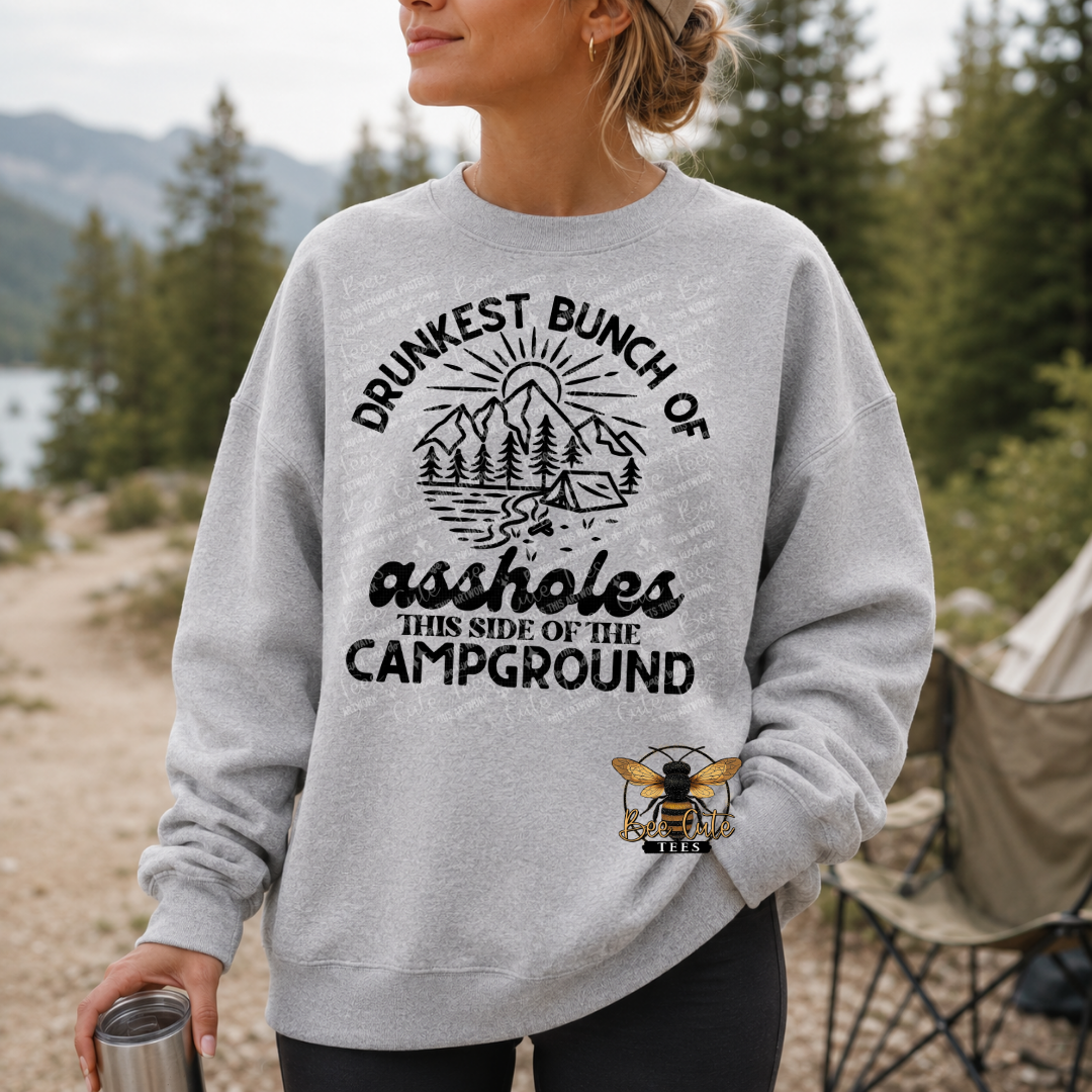 A woman wearing a light gray crewneck sweatshirt with a black graphic that reads 'DRUNKEST BUNCH OF assholes THIS SIDE OF THE CAMPGROUND' with a mountain and tent illustration, and a bee logo near the bottom. The background shows an outdoor camping scene with trees, a tent, and a folding chair.