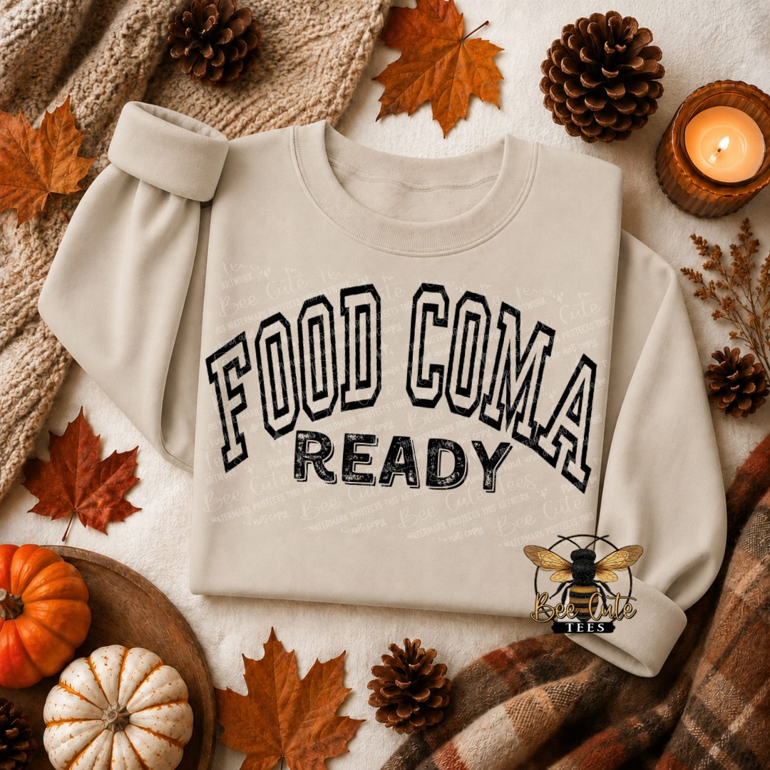 A cream-colored sweatshirt with 'FOOD COMA READY' printed on the front, styled in a cozy autumn flat-lay with pinecones, fall leaves, a candle, and a plaid blanket.