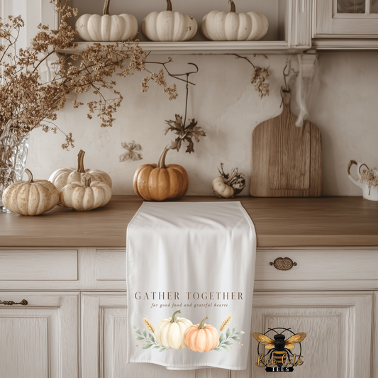 A white kitchen tea towel draped over a wooden countertop in a fall-themed kitchen. The runner features the text 'GATHER TOGETHER for good food and great company' above a watercolor illustration of three pumpkins in beige, white, and orange, with wheat ears.