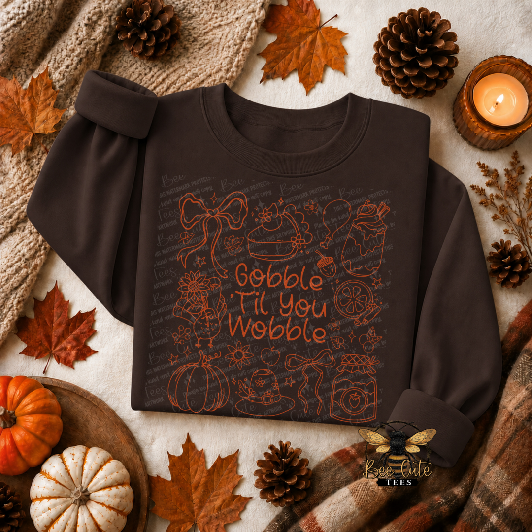 A dark brown long-sleeve sweatshirt laid flat on a cream fabric background with autumn props including pinecones, maple leaves, pumpkins, a knitted blanket, and a lit candle. The sweatshirt has a large front graphic with orange line drawings and the phrase 'Gobble Til You Wobble' in orange lettering.