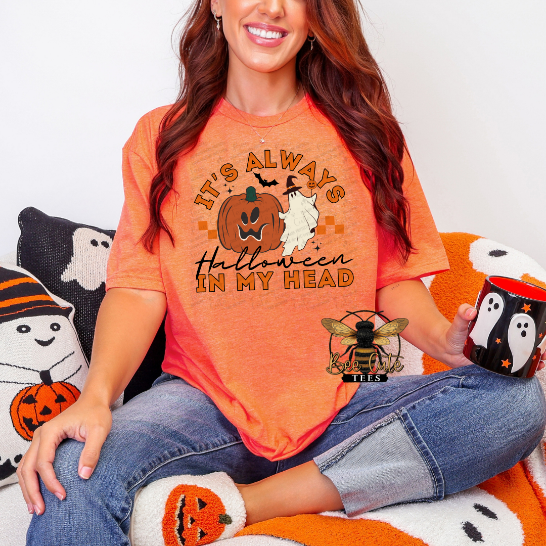 A woman wearing a bright orange graphic t-shirt with a Halloween design that reads 'IT'S ALWAYS Halloween IN MY HEAD' with a jack-o'-lantern, ghost, and bat. She is sitting on a cozy orange and white blanket with Halloween-themed pillows and holding a black mug with a ghost design.
