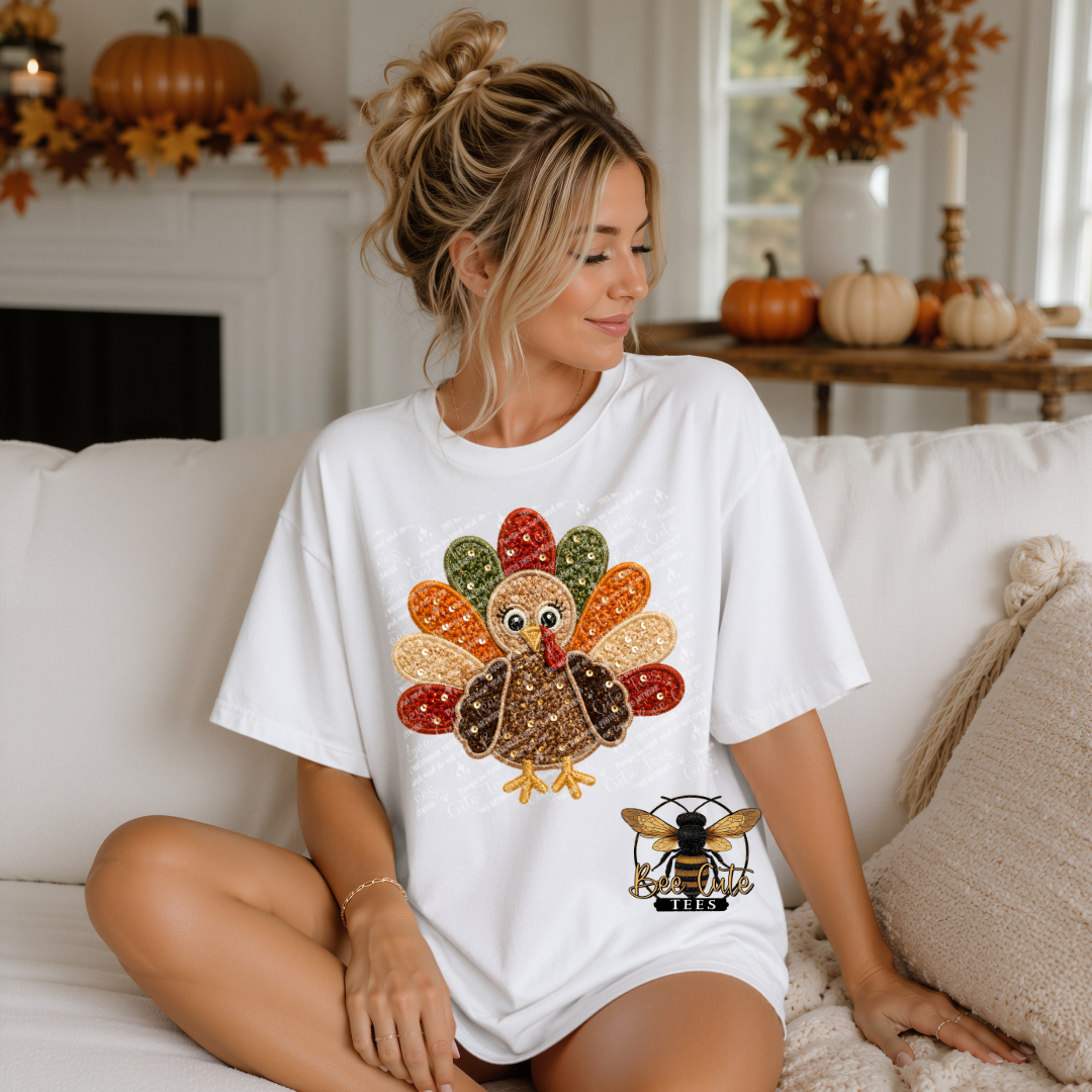 A woman sits on a white couch wearing an oversized white graphic T-shirt with a colorful turkey design, in a cozy fall-themed living room setting.
