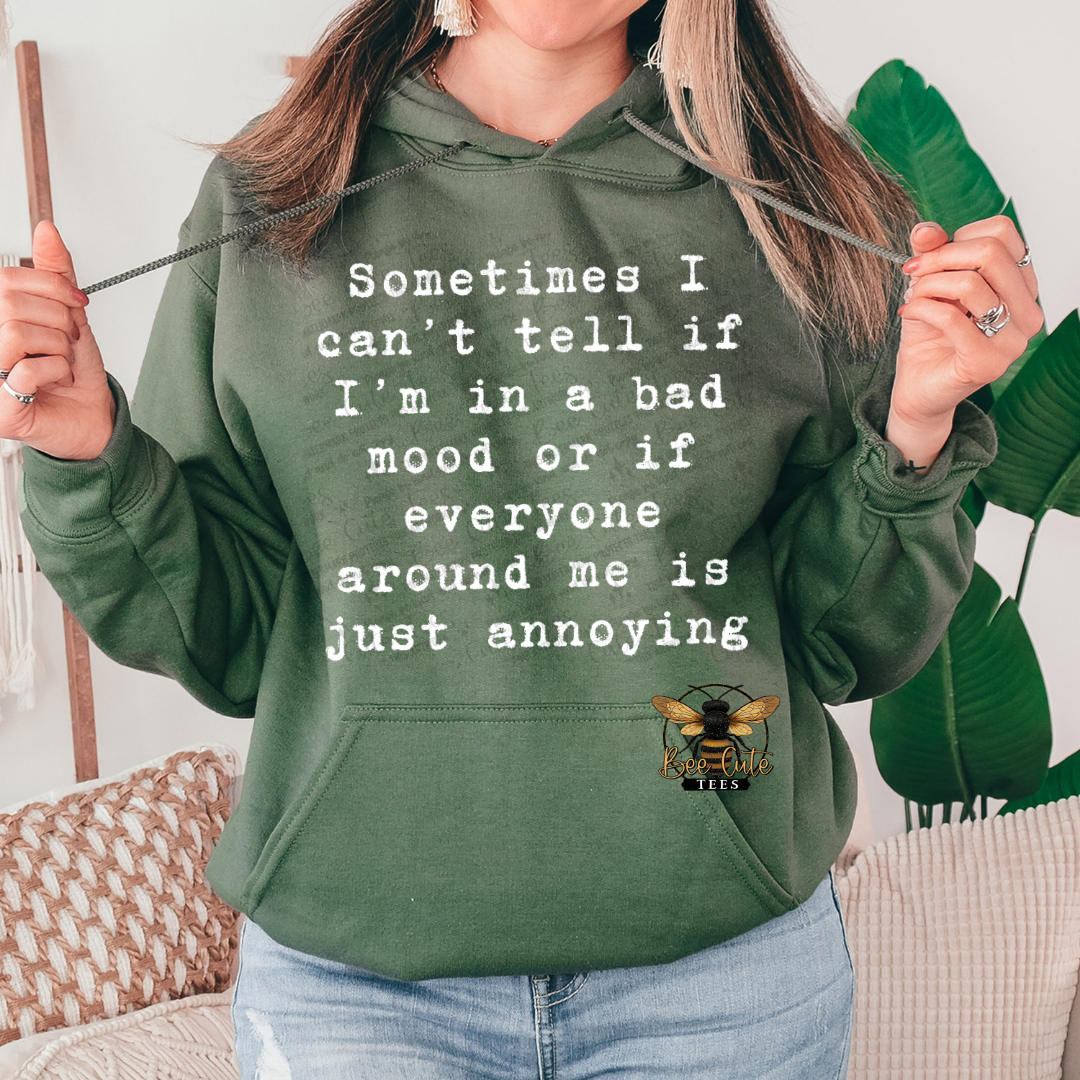 A woman wearing an olive green hoodie with white text that reads 'Sometimes I can't tell if I'm in a bad mood or if everyone around me is just annoying' and a small bee logo near the pocket. She is holding the drawstrings of the hoodie, with a couch and green leaves in the background.