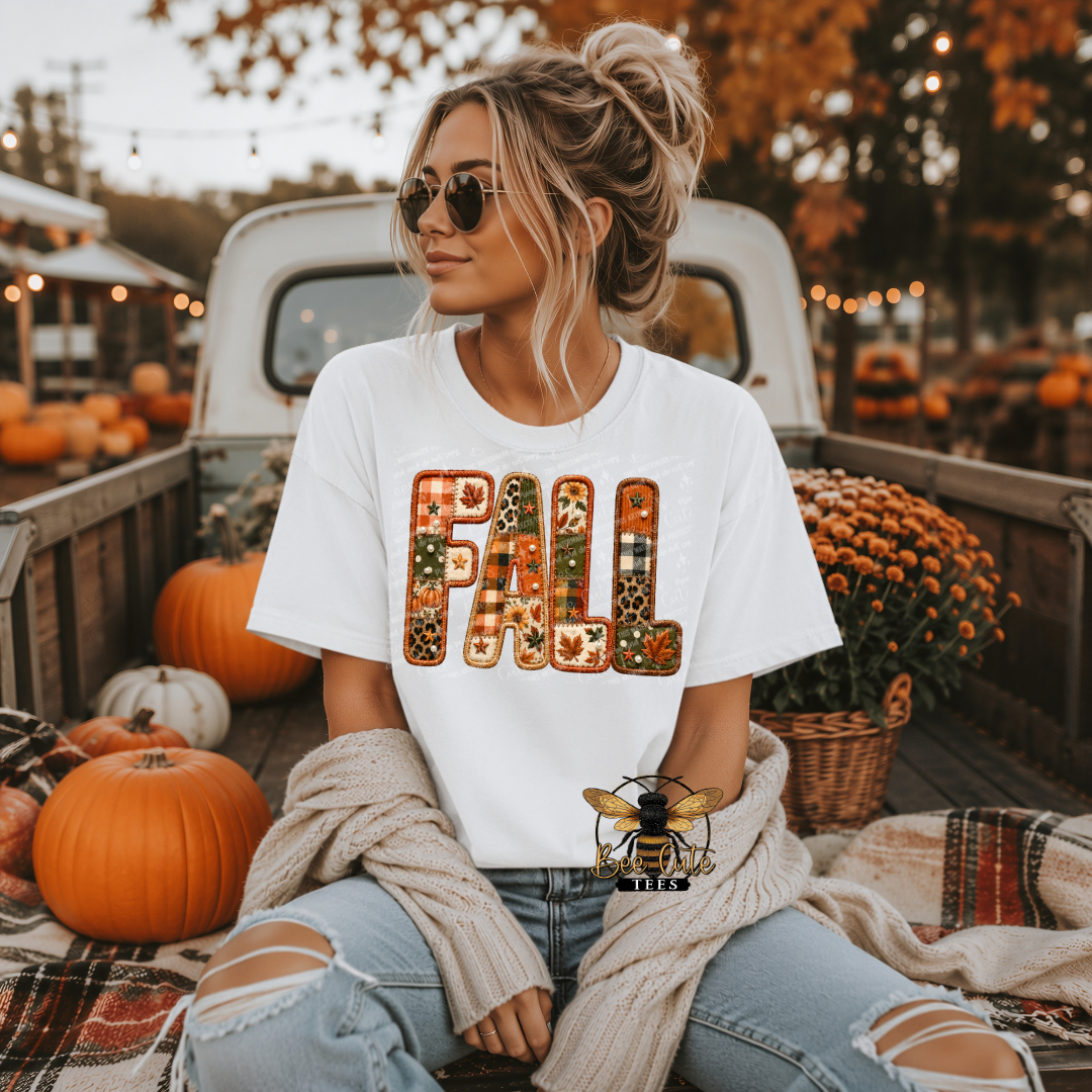 A woman sitting in the bed of a pickup truck surrounded by pumpkins and fall decorations, wearing a white graphic t-shirt with the word 'FALL' filled with autumn-themed patterns.