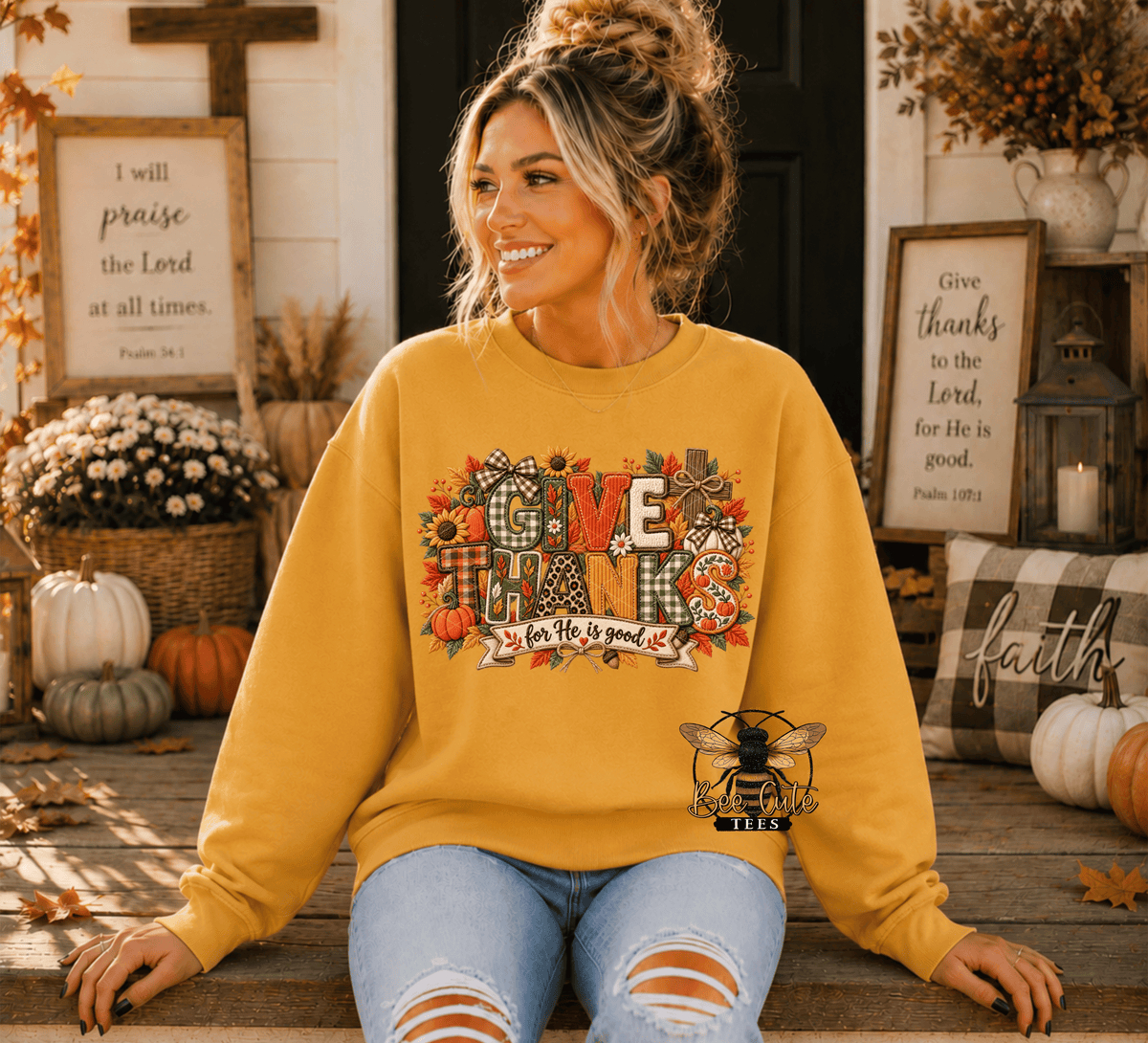 A woman wearing a mustard yellow crewneck sweatshirt with a colorful 'GIVE THANKS' autumn graphic sits on a wooden porch. The graphic features pumpkins, fall leaves, and a bee logo.