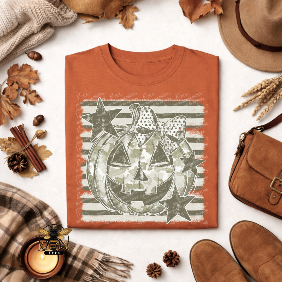 An orange crewneck t-shirt with a distressed graphic of a jack-o'-lantern, stars, and a bow on a striped background, styled in an autumn flat-lay with a hat, scarf, shoes, and candle.