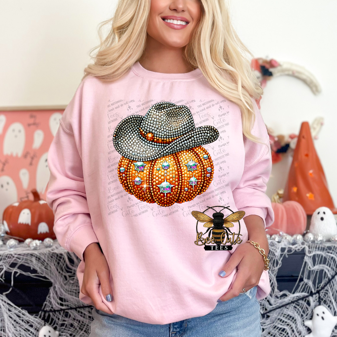 A woman wearing a light pink sweatshirt with a large Halloween-themed graphic featuring a rhinestone-embellished cowboy hat and pumpkin, posed in a decorated indoor setting with autumn and Halloween decorations.