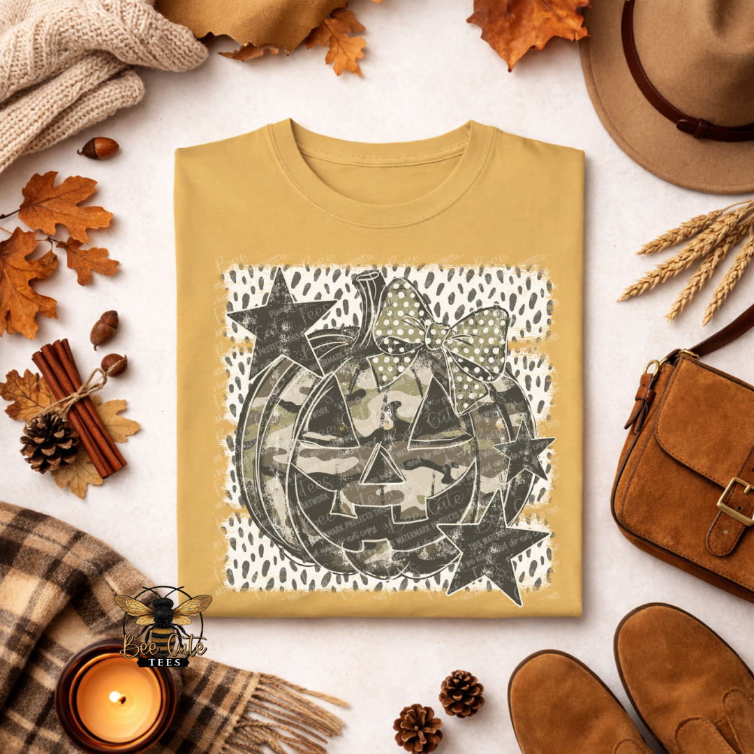 A folded mustard crewneck sweatshirt with a graphic print of a jack-o'-lantern made of camouflage patterns, stars, and a bow, styled in an autumn flat-lay with a hat, shoes, scarf, candle, and leaves.