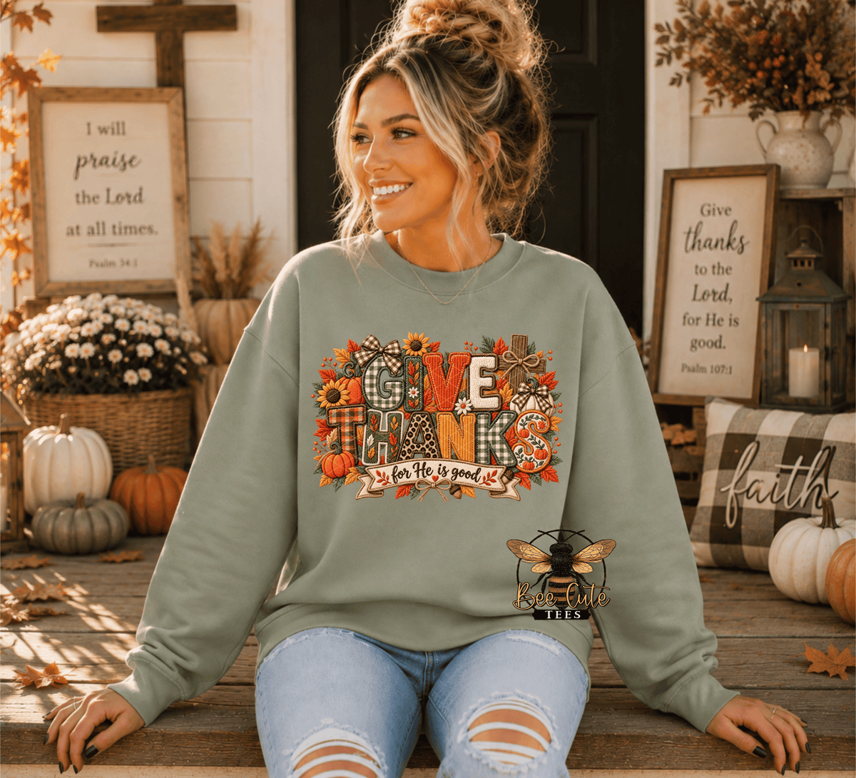 A woman wearing a sage green crewneck sweatshirt with a colorful 'GIVE THANKS' autumn graphic, sitting on a wooden porch with fall decorations like pumpkins and framed signs.