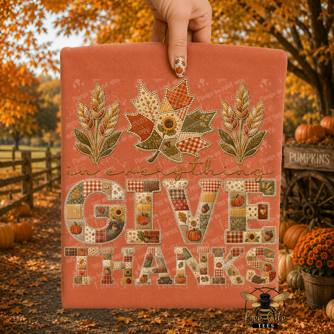 A hand holds a terracotta-colored folded t-shirt with a patchwork autumn design that reads 'in everything GIVE THANKS.' The sign features three leaf motifs at the top and decorative lettering filled with small patchwork squares.
