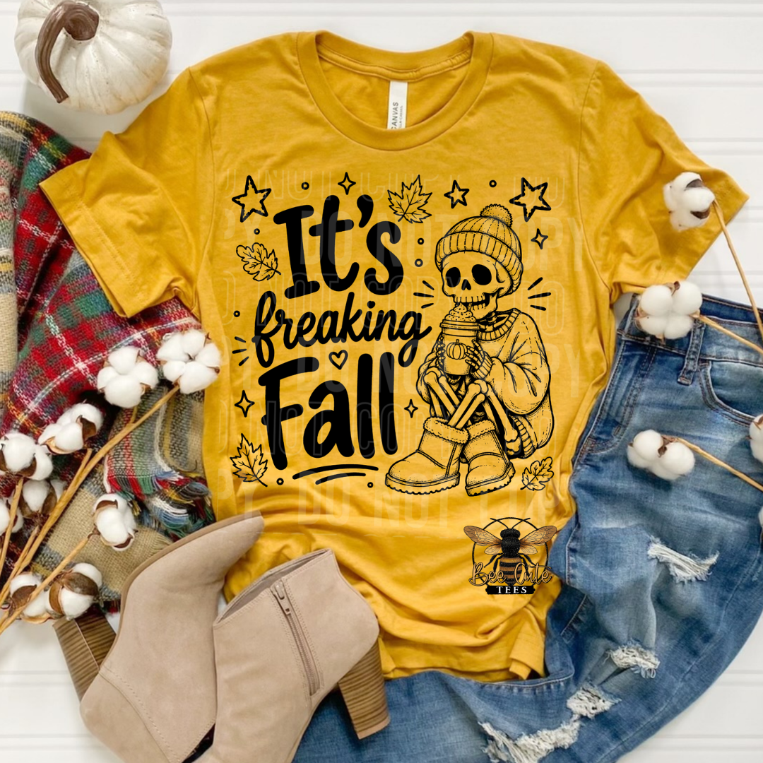 A yellow graphic T-shirt laid flat with autumn styling props including blue jeans, beige ankle boots, a plaid scarf, cotton bolls, and a small pumpkin. The shirt reads “It’s Breaking Fall” with a skeleton wearing a beanie and holding a mug, surrounded by stars and leaves.
