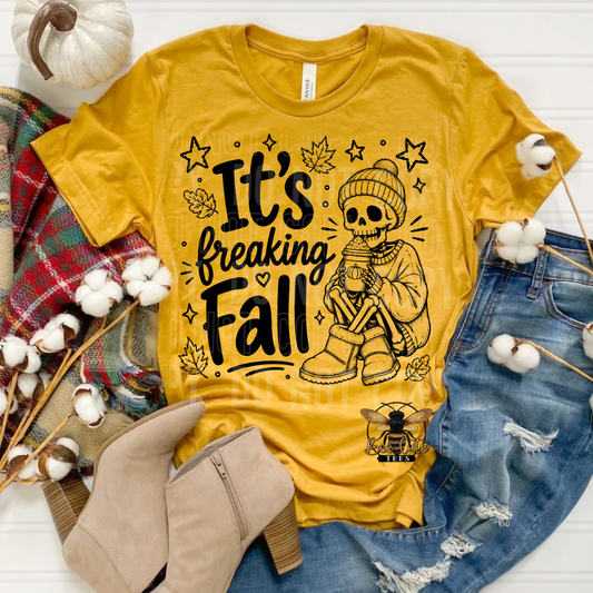 A yellow graphic T-shirt laid flat with autumn styling props including blue jeans, beige ankle boots, a plaid scarf, cotton bolls, and a small pumpkin. The shirt reads “It’s Breaking Fall” with a skeleton wearing a beanie and holding a mug, surrounded by stars and leaves.