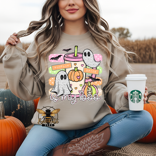 A woman sitting outdoors among pumpkins wears a beige graphic sweatshirt with a Halloween-themed print featuring ghosts, pumpkins, a drink, and the phrase 'is my boo'. She holds a white Starbucks coffee cup in one hand and wears blue jeans and brown cowboy boots.