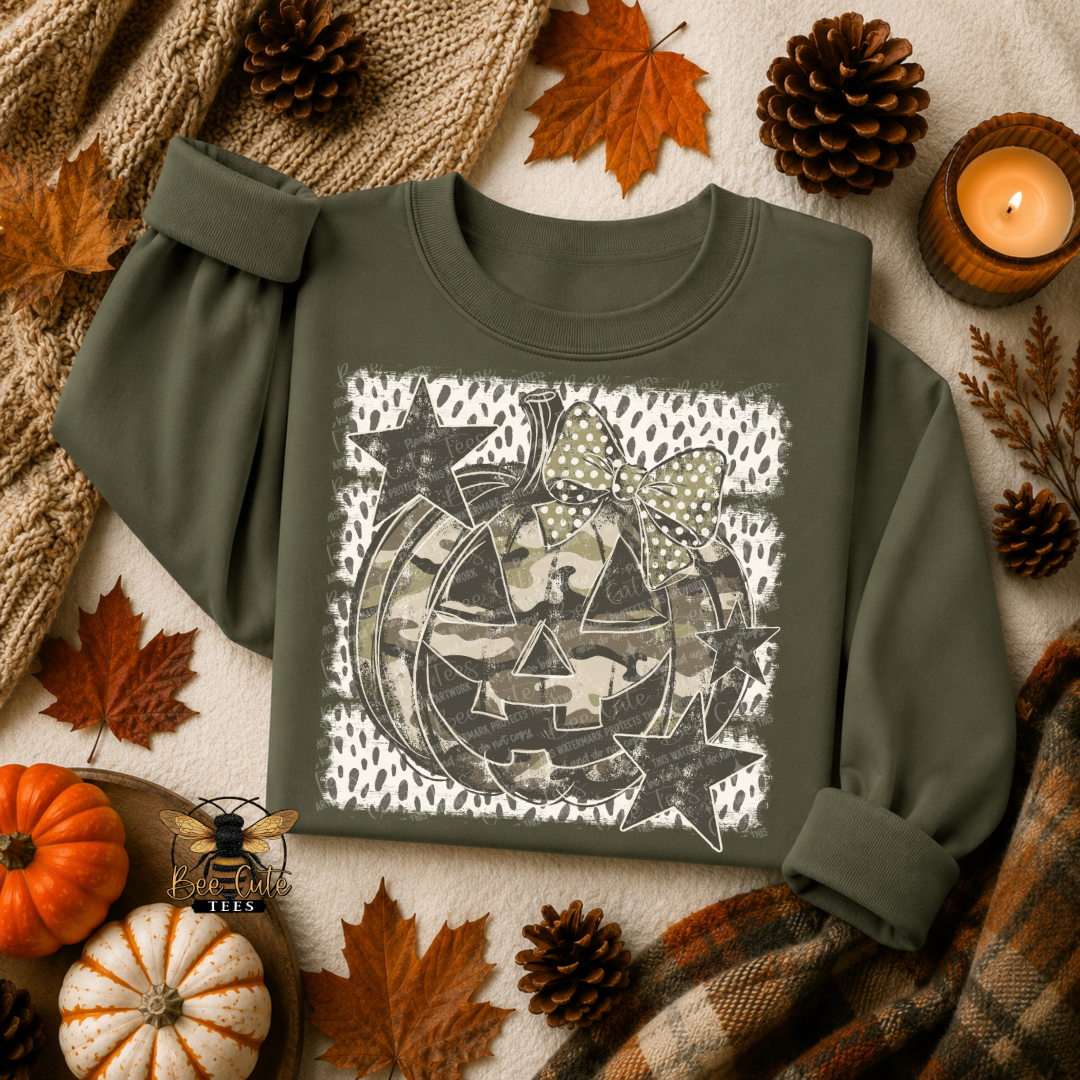 An olive green long-sleeve t-shirt with a Halloween-themed graphic print featuring a jack-o'-lantern, stars, and a bow, styled in a flat-lay autumn setting with pumpkins, pinecones, and a candle.