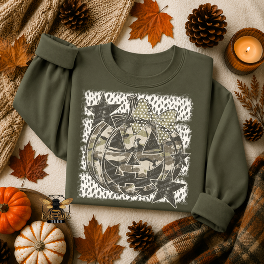 An olive green long-sleeve t-shirt with a Halloween-themed graphic print featuring a jack-o'-lantern, stars, and a bow, styled in a flat-lay autumn setting with pumpkins, pinecones, and a candle.