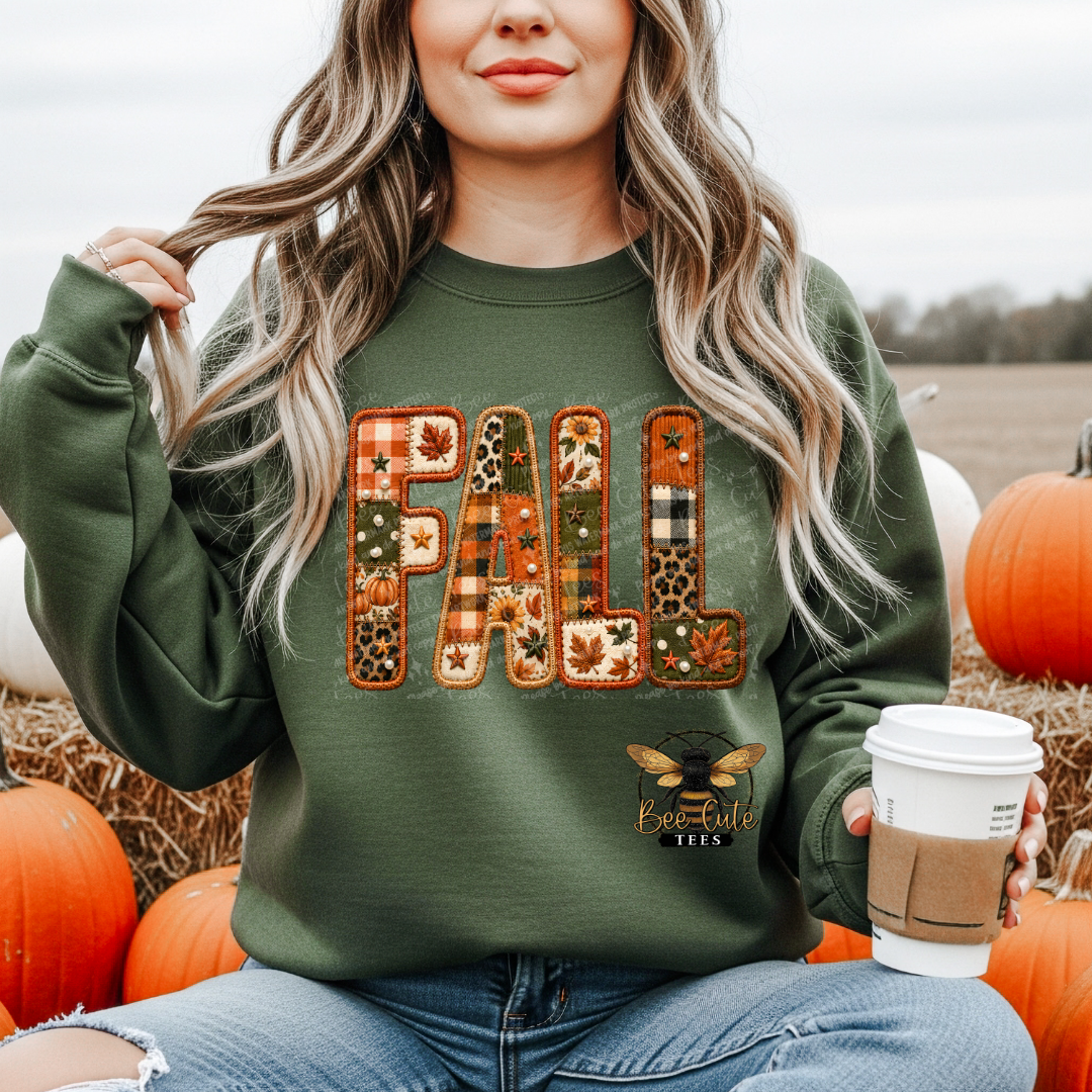 A woman wearing an olive green crewneck sweatshirt with a large 'FALL' graphic made of autumn-themed patchwork patterns, sitting outdoors among pumpkins and hay.
