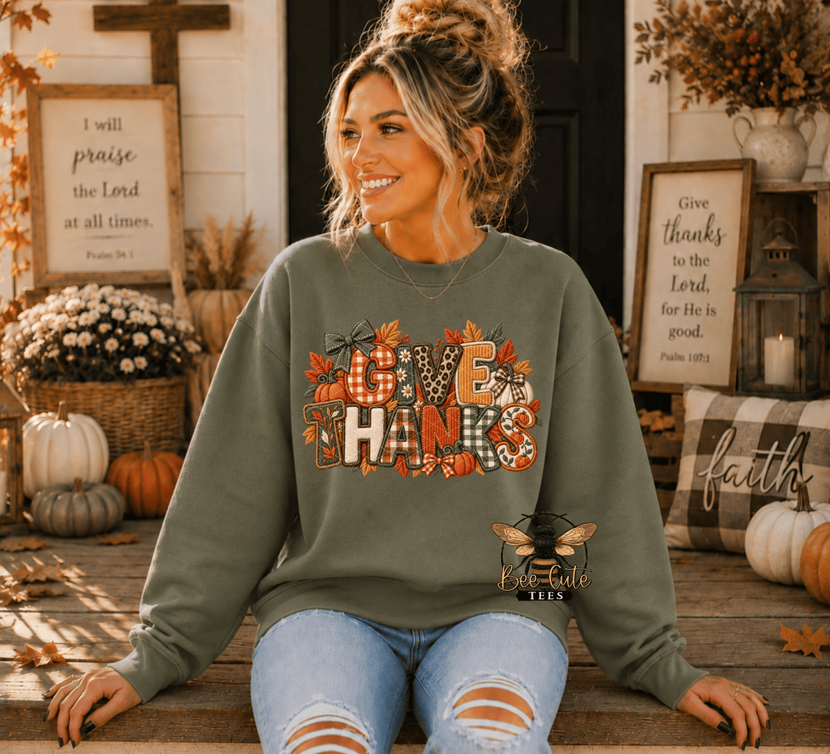 A woman wearing an olive green crewneck sweatshirt with a 'GIVE THANKS' autumn graphic sits on a wooden porch decorated for Thanksgiving, with framed scripture and pumpkins in the background.