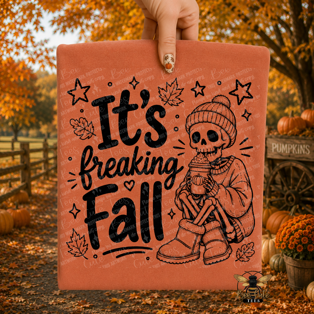 A hand holds up a terracotta-colored folded t-shirt featuring a black graphic of a skeleton sitting in a beanie and holding a coffee cup, surrounded by the text 'It's freaking Fall' with autumn leaves and stars. The background is a blurred outdoor fall scene with pumpkins, orange foliage, and a wooden fence.