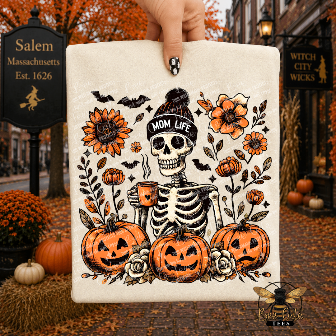 A hand holds a beige folded t-shirt featuring a Halloween-themed illustration of a skeleton wearing a 'MOM LIFE' beanie, surrounded by pumpkins, flowers, and bats. The background shows an autumn street scene with Salem, Massachusetts street signs and Halloween decorations.