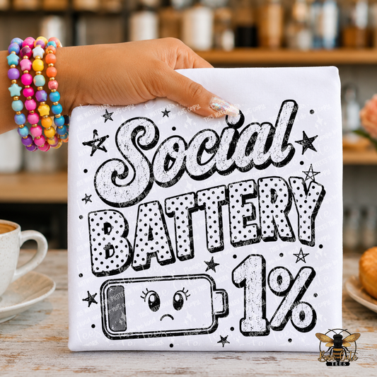 A hand wearing colorful beaded bracelets holds a white  T-shirt-style graphic that reads “Social Battery 1%” in black retro lettering with stars and a sad battery illustration. The background shows a blurred cafe interior with a cup, table surface, and pastries.