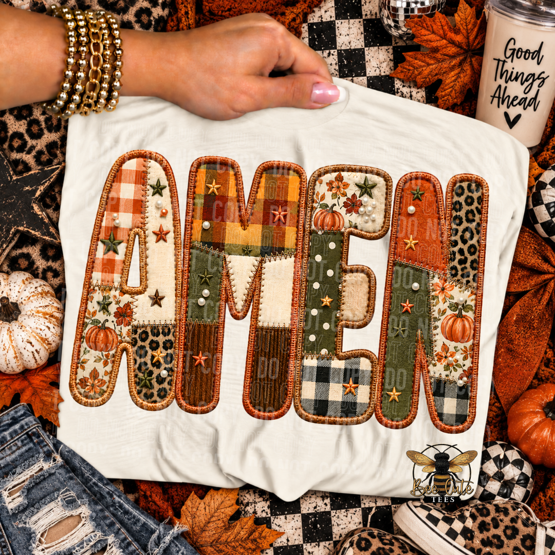 A white graphic T-shirt laid flat with a hand holding the collar, surrounded by autumn props including pumpkins, leaves, a checkered blanket, denim, bracelets, and a coffee cup. The shirt features large patchwork-style letters spelling 'AMEN' in orange, brown, green, beige, and black patterns.