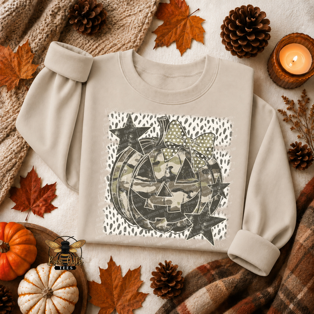 A beige long-sleeve sweatshirt laid flat on a cozy autumn-themed background with pinecones, fall leaves, a candle, pumpkins, and plaid fabric. The sweatshirt has a square front graphic featuring a jack-o'-lantern face made from camouflage-patterned stars and stripes, with a polka-dot bow above it.