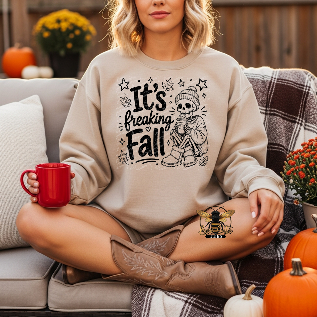 A woman sits cross-legged on a couch wearing a beige crewneck sweatshirt with a black graphic that reads 'It's Breaking Fall' alongside a skeleton illustration. She is holding a red mug, with pumpkins, a plaid blanket, and autumn flowers in the background.