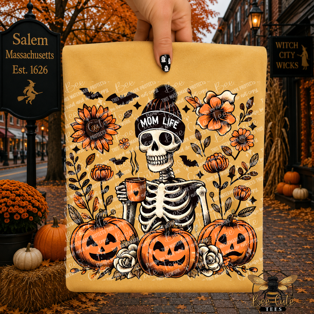 A hand holds a yellow folded t-shirt featuring a Halloween-themed skeleton illustration with jack-o'-lanterns, flowers, bats, and a beanie labeled 'MOM LIFE.' The background shows a real outdoor street scene with autumn leaves, pumpkins, and storefront signs.
