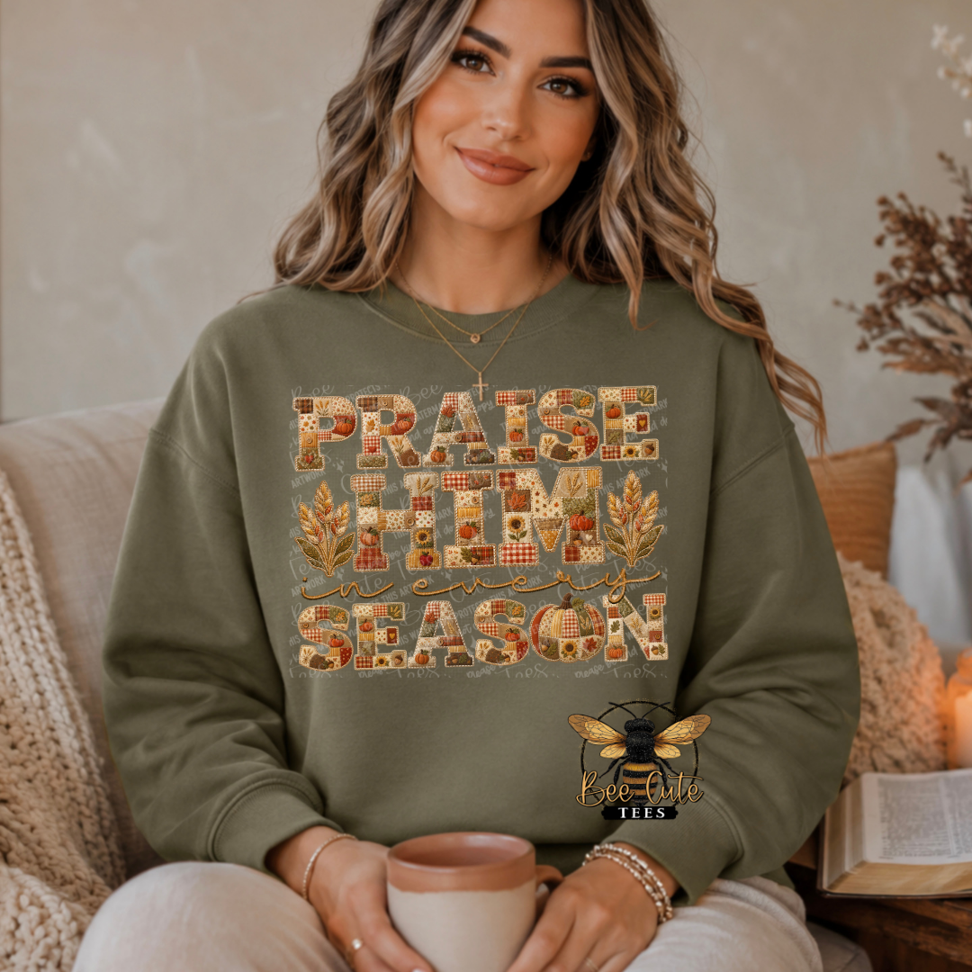 A woman wearing an olive green sweatshirt with a 'Praise Him Season' graphic made of autumn leaves and patterned squares, holding a mug while sitting in a cozy indoor setting.