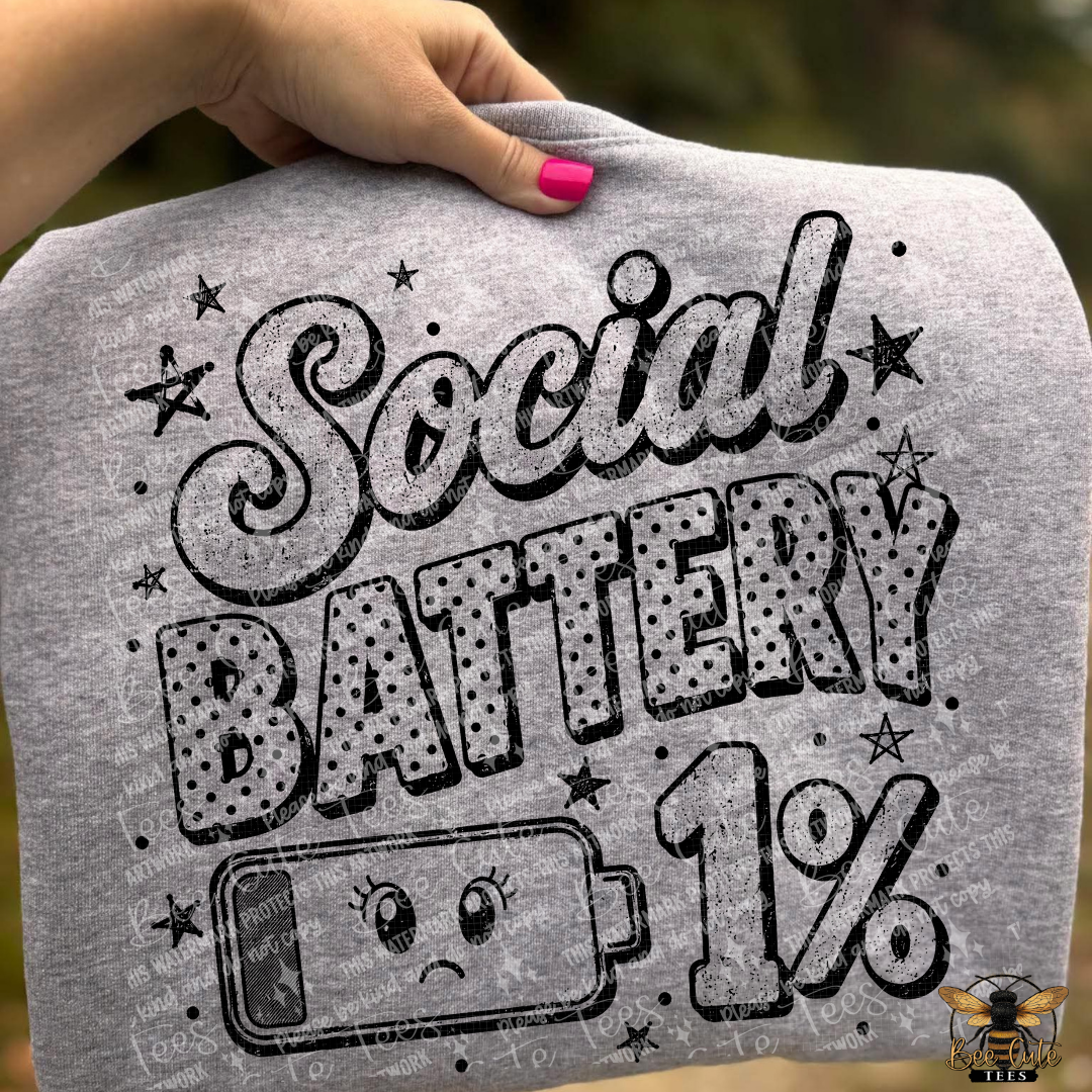 A person's hand with pink nail polish holds up a heather gray t-shirt featuring a black graphic that reads 'Social Battery 1%' with a sad battery illustration.