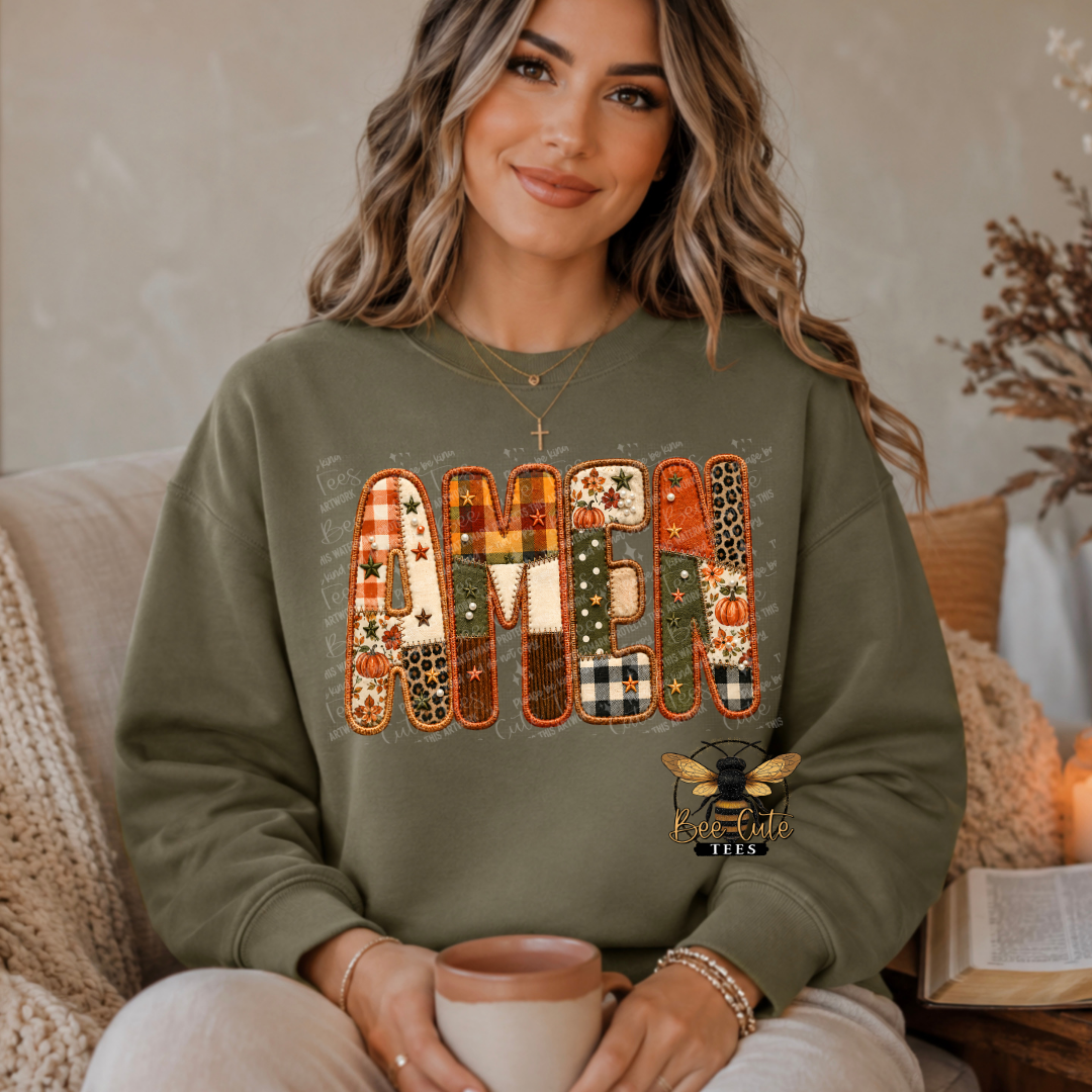 A woman wearing an olive green sweatshirt with a large 'AMEN' patchwork graphic in autumn colors, sitting in a cozy indoor setting and holding a mug.