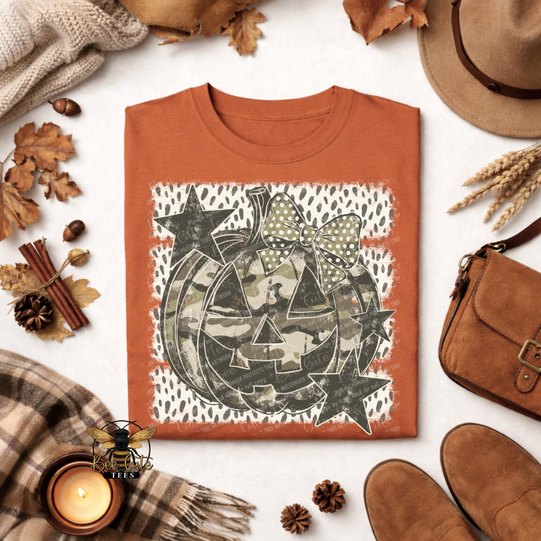 An orange crewneck t-shirt with a Halloween-themed graphic print of a jack-o'-lantern in camouflage, surrounded by autumn props like a hat, scarf, candle, and shoes on a white background.