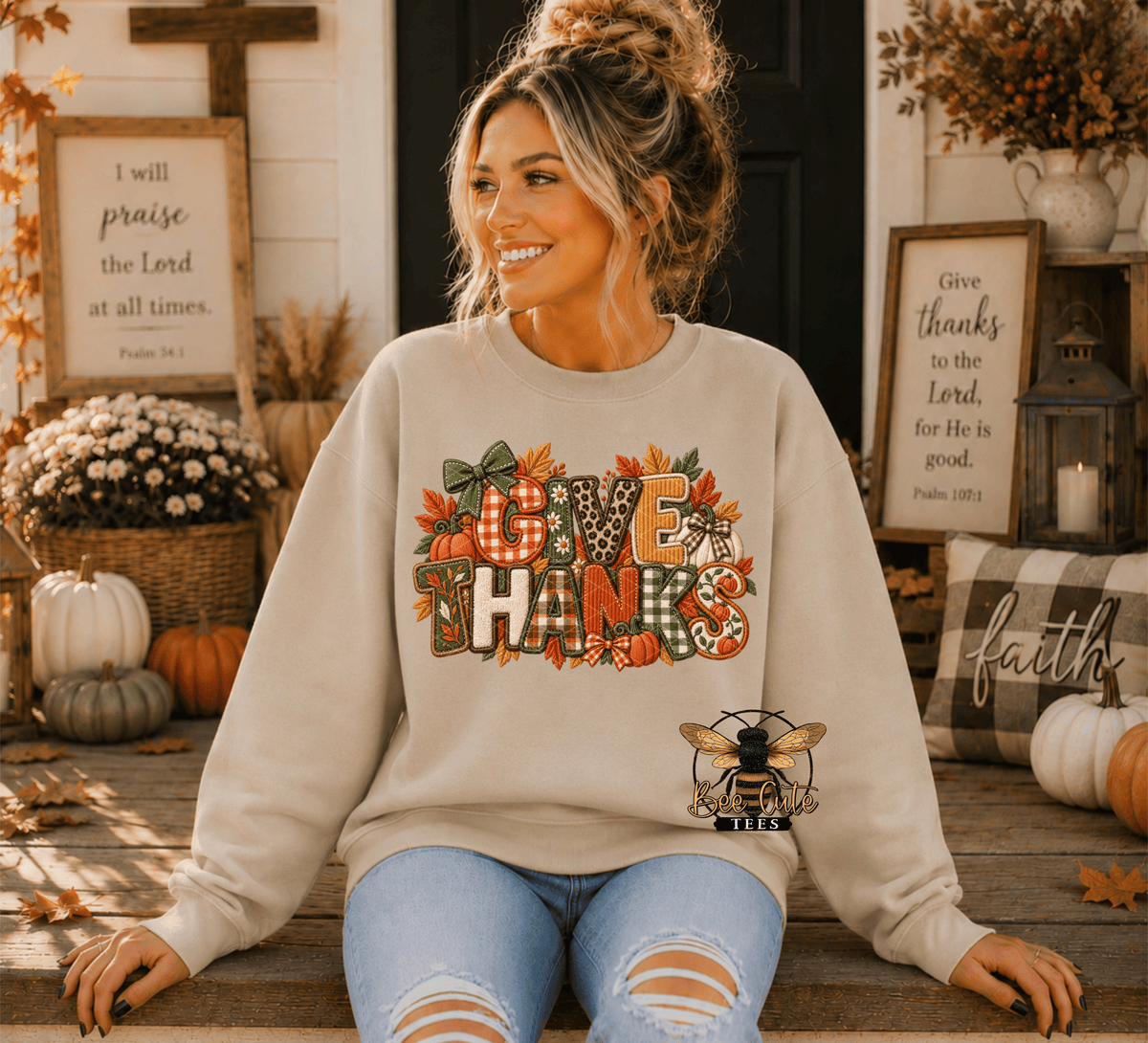 A woman wearing an beige crewneck sweatshirt with a 'GIVE THANKS' autumn graphic sits on a wooden porch decorated for Thanksgiving, with framed scripture and pumpkins in the background.