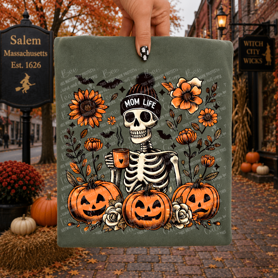 A hand holds a green folded t-shirt featuring a Halloween-themed skeleton holding a mug, surrounded by pumpkins, flowers, bats, and the text 'MOM LIFE.' The background shows an outdoor autumn street scene with fall leaves, lanterns, and a Salem street sign.