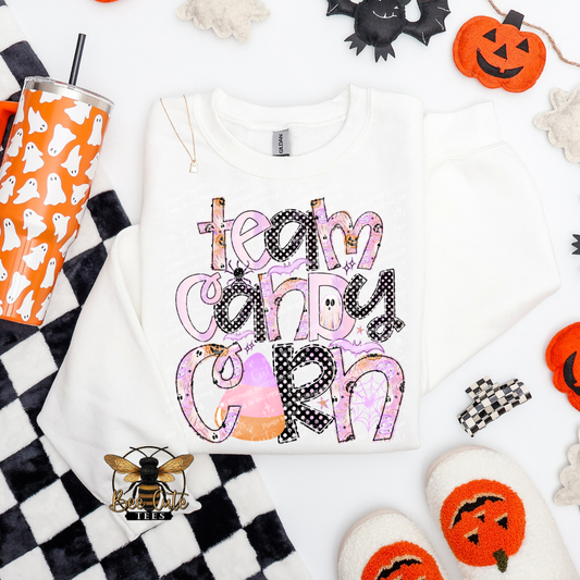 A white long-sleeve crewneck sweatshirt with a colorful 'team candy corn' graphic, styled in a flat lay with Halloween props including a ghost cup, jack-o'-lanterns, and a checkered blanket.