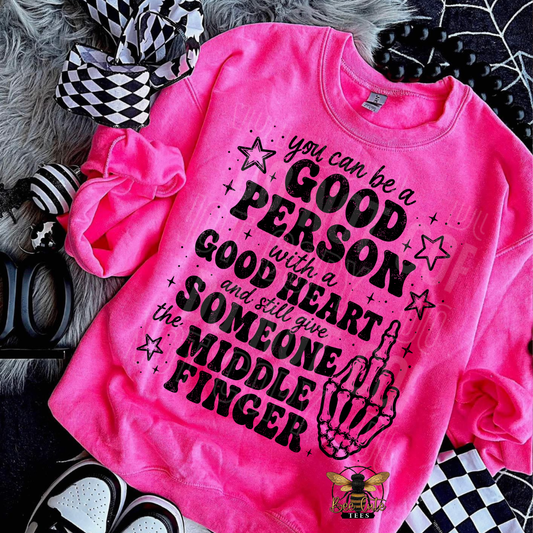 A bright pink crewneck sweatshirt with a bold black graphic text that reads 'you can be a GOOD PERSON with a GOOD HEART and still give SOMEONE the MIDDLE FINGER' along with a skeletal hand graphic. The sweatshirt is styled in a flat lay with a black and white checkered bow, a black scrunchie, and black-and-white sneakers on a gray faux fur background.