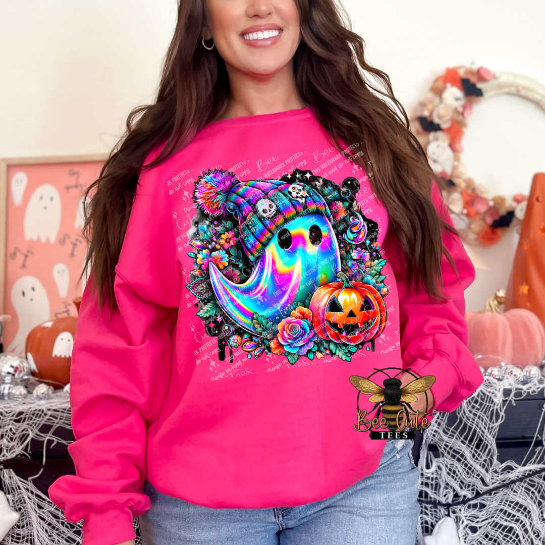 A woman wearing a bright pink sweatshirt with a colorful Halloween graphic featuring a rainbow ghost, flowers, a pumpkin, and a hat. The sweatshirt has a small bee logo on the lower right.