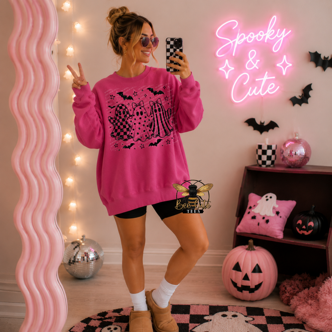 A woman takes a mirror selfie wearing an oversized bright pink sweatshirt with a black ghost-themed graphic, paired with black shorts, white socks, and tan boots. The room is decorated for Halloween with a pink neon sign reading 'Spooky & Cute', a pink wavy mirror, pumpkins, and a pink ghost pillow.