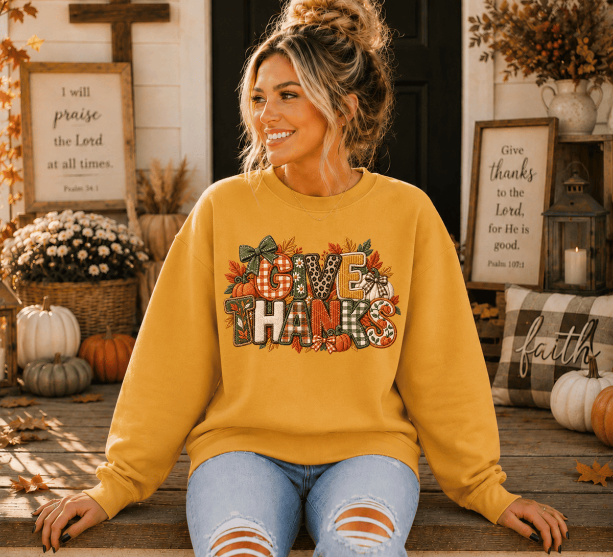 A woman wearing a mustard yellow crewneck sweatshirt with a colorful 'GIVE THANKS' graphic featuring autumn leaves, pumpkins, and plaid patterns, sitting on a wooden porch surrounded by fall decorations.