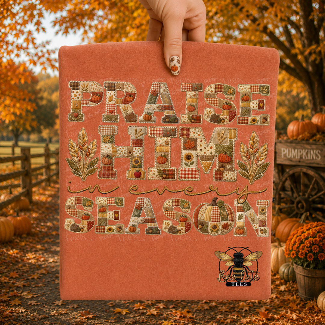 A hand holds up a terracotta-colored folded t-shirt featuring a patchwork-style autumn design with the text 'PRAISE HIM IN EVERY SEASON' and a bee logo. The background shows an outdoor fall scene with pumpkins, orange foliage, and a wooden fence.