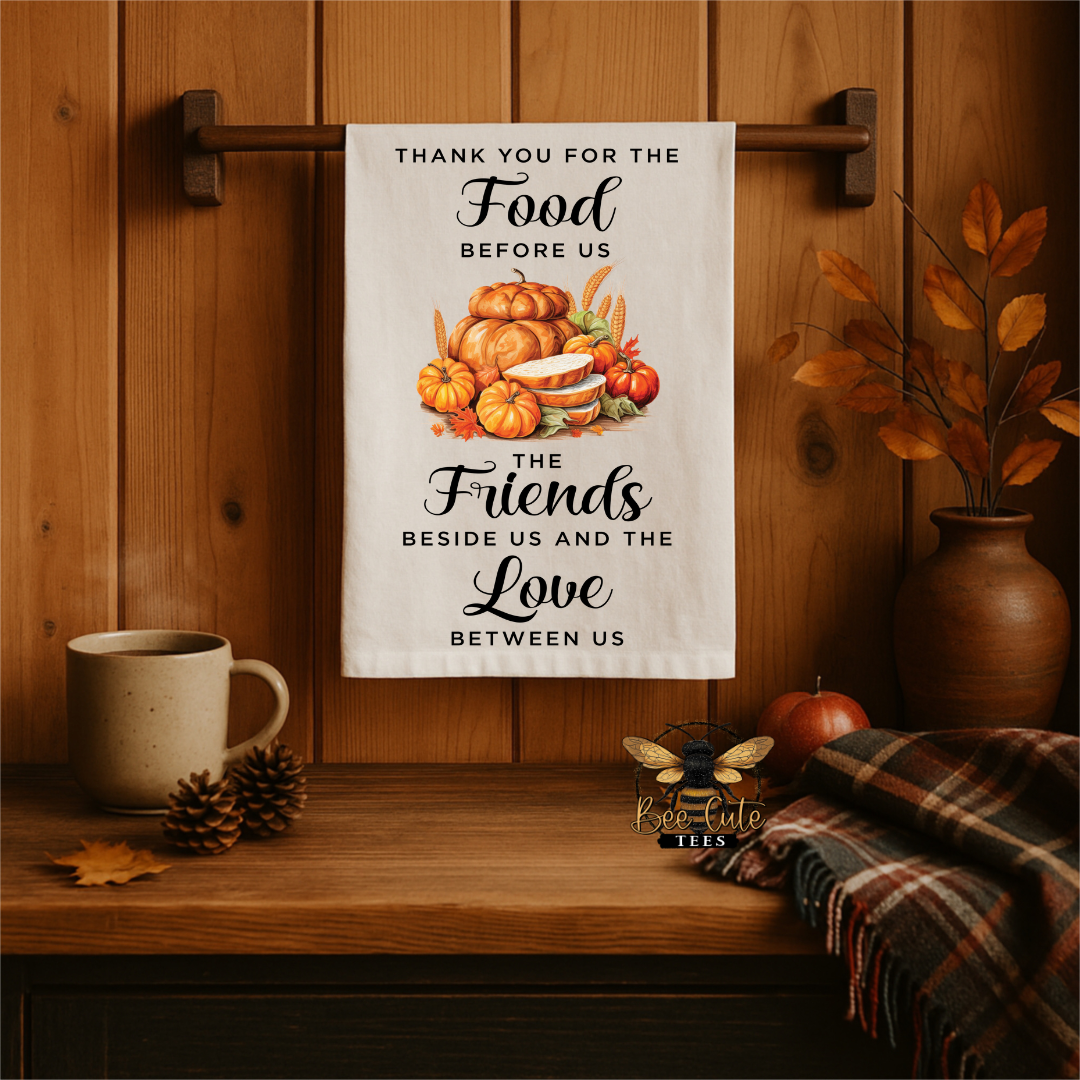 A white kitchen towel with a fall harvest illustration and the text 'THANK YOU FOR THE Food BEFORE US THE Friends BESIDE US AND THE Love BETWEEN US' hangs from a wooden rack on a wood-paneled wall. The scene is styled with a mug, pinecones, a plaid throw, and a vase of autumn leaves.