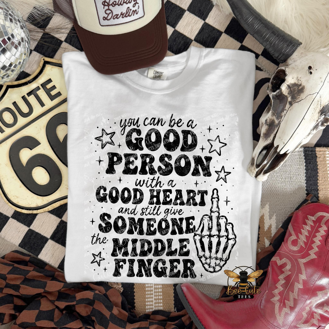 A white graphic t-shirt laid flat on a western-themed background with a brown trucker hat, a Route 66 sign, a skull, and red cowboy boots. The shirt features a black text graphic that reads 'you can be a GOOD PERSON with a GOOD HEART and still give SOMEONE the MIDDLE FINGER' alongside a hand graphic.
