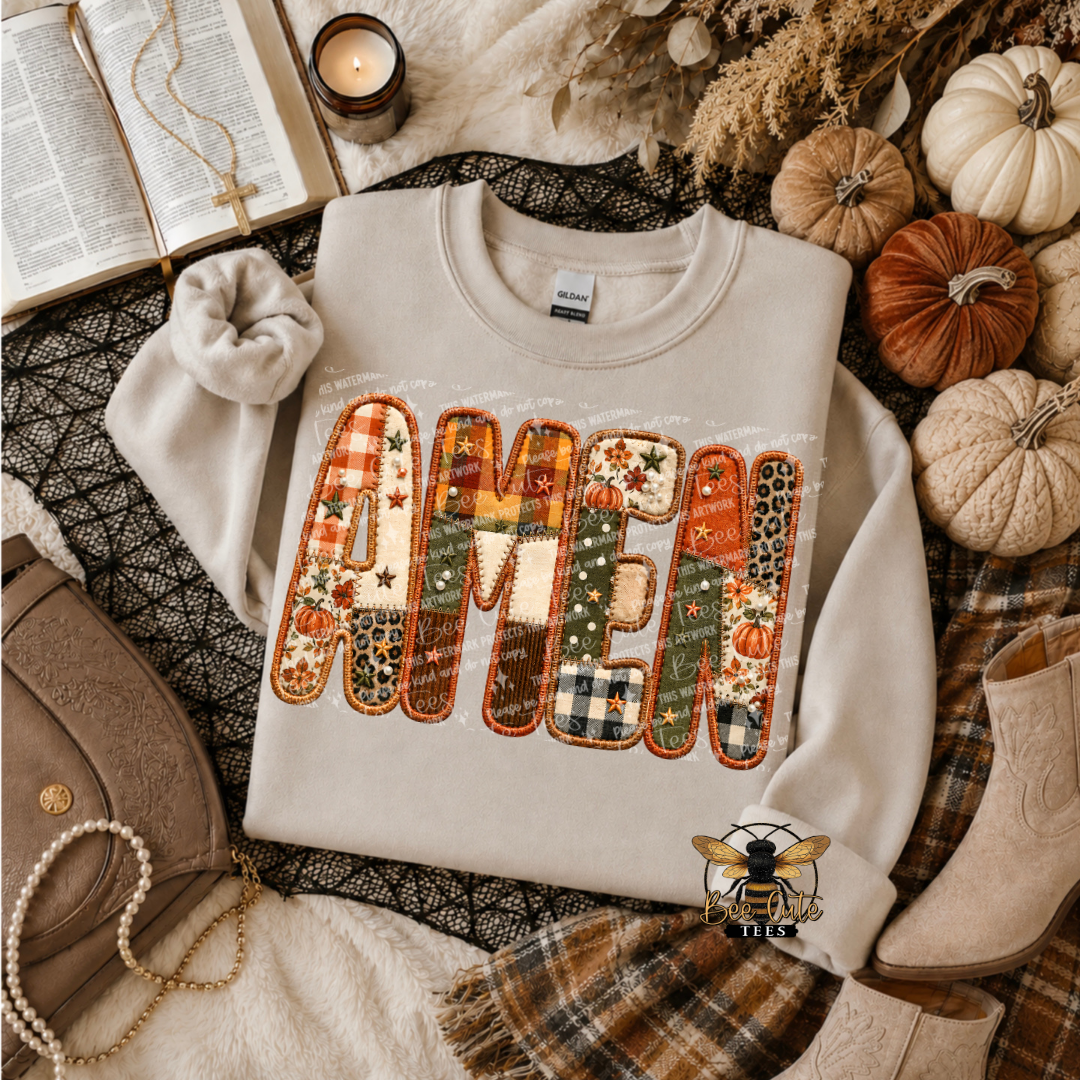 A light beige crewneck sweatshirt laid flat in a styled autumn scene, featuring the word 'AMEN' in large patchwork letters with fall patterns like plaid, leopard print, and pumpkins. The sweatshirt is surrounded by pumpkins, a book, a candle, a small purse, and boots.