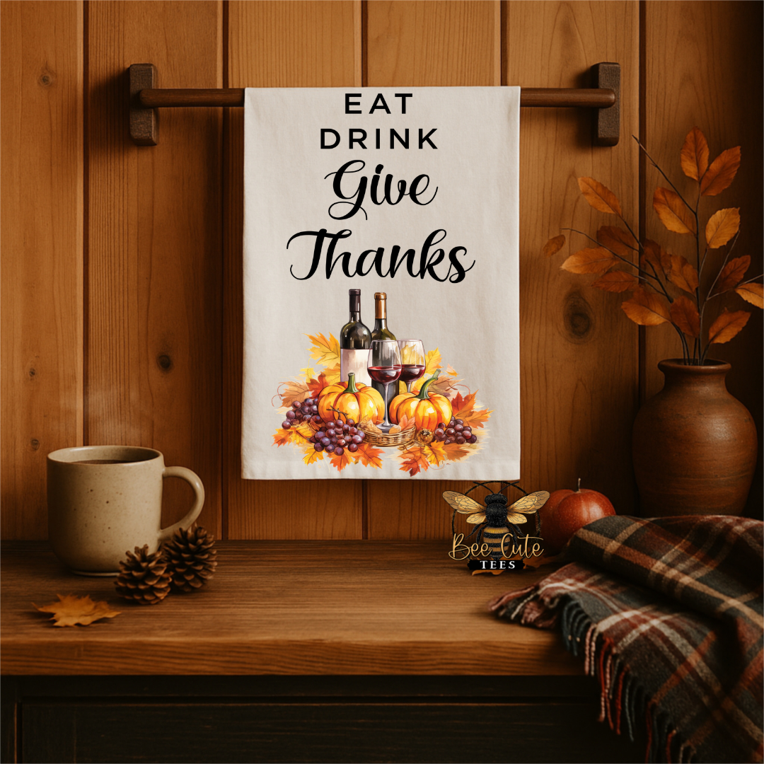 A white kitchen towel with the text 'EAT DRINK Give Thanks' and a fall harvest illustration of pumpkins, grapes, and wine bottles, hanging on a wooden wall rack in a styled autumn kitchen scene.